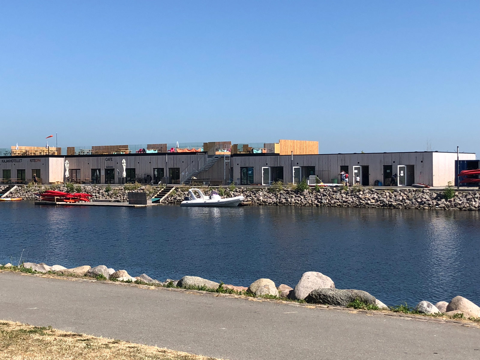 Kajakker og både, som lægger i vandet ved Amager Strand. Oppe på land står Kajakhotellet med træbeklædning. på taget er tagterrassen med parasoller.