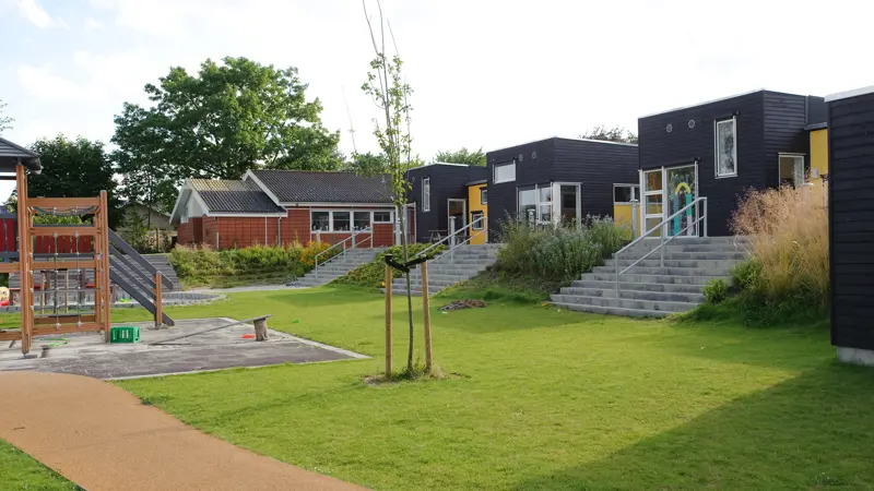 Børneinstitution Grønnemarken udefra - den forskudte facade med forskellige rum. En legeplads i en stor sandkasse træer og trapper op til børnehavens indgang