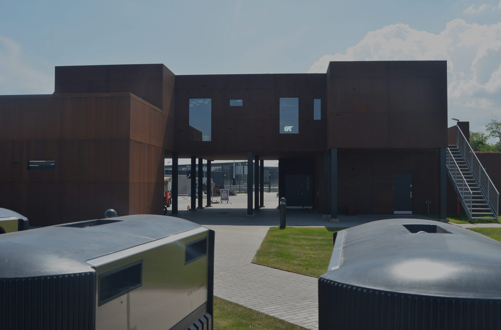 2 store skraldespande i front, en gangsti hen til bygningen i cortens stål. Mørk overflade