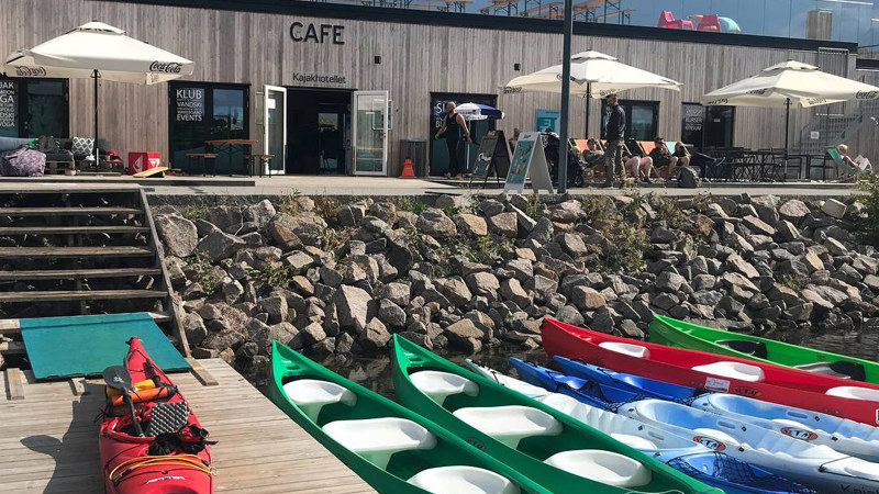 Kajakker i vandkanten og en rød på broen. Der er en trappe op mod Kajakhotellet hvor parasollerne er slået op man kan se borde og stole på tagterrassen.