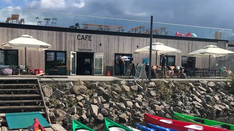 Kajakker i vandkanten og en rød på broen. Der er en trappe op mod Kajakhotellet hvor parasollerne er slået op man kan se borde og stole på tagterrassen.
